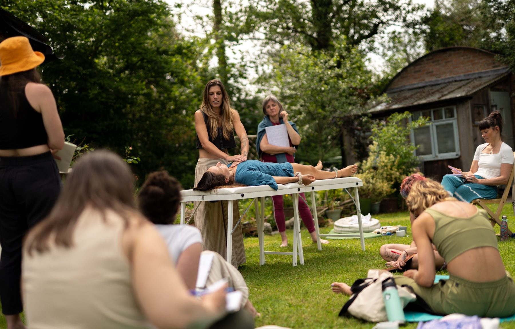 Madison demonstrating on a client lying on a table in a garden, with a group of trainees watching and taking notes